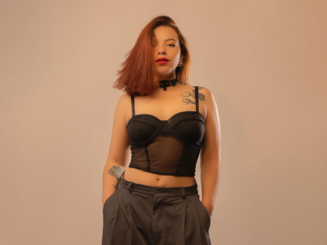 SabriinaMorgan with red hair, black sheer bra, black choker, black pants, hands in pockets, tattoos on arm and chest, standing, looking at camera, medium shot, sexy.