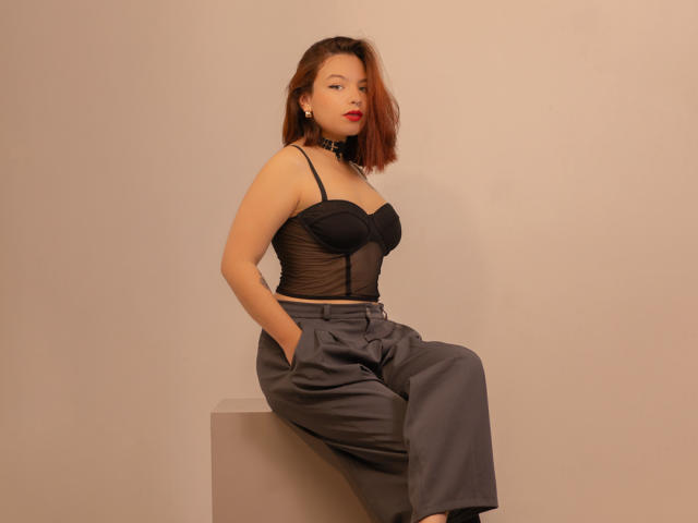 SabriinaMorgan with red lipstick, black sheer top, and gray pants, sitting on a cube, hands in pockets, looking at camera, medium skin, brown bob, sexy, confident.