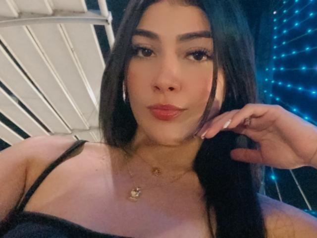 DayanaWatson with long black hair, wearing a black spaghetti strap top, has a cute face, full pink lips, and is touching her hair with a gold necklace.