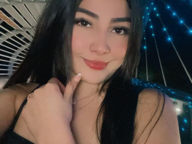 DayanaWatson has long black hair, dark eyebrows, red lipstick, and a cute nose ring. She's smiling with her finger touching her cheek, wearing a black top.