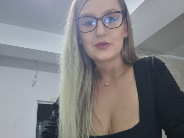 MayaTrainor with long blonde hair, glasses, and red lipstick, wearing a black top, showing her tits in a selfie, slightly pouting her plump, pretty pink lips.
