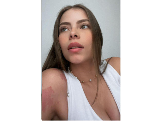 AnnaFer, avec de longs cheveux bruns, un débardeur blanc, un petit collier en or, et une légère ecchymose sur son épaule droite, regardant sur le côté avec des lèvres roses..