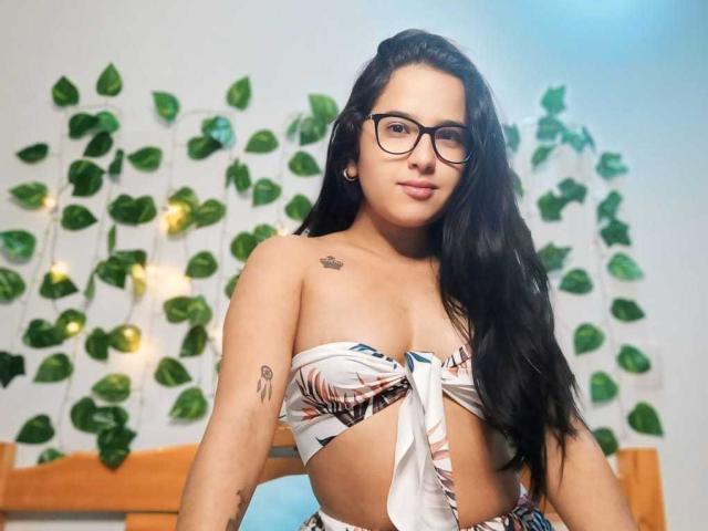 AmeliaLea met lang zwart haar, een bril, en een opgetrokken, gestreept bikini top, die haar borsten en tatoeages laat zien, kijkt naar de camera met een lichte glimlach..