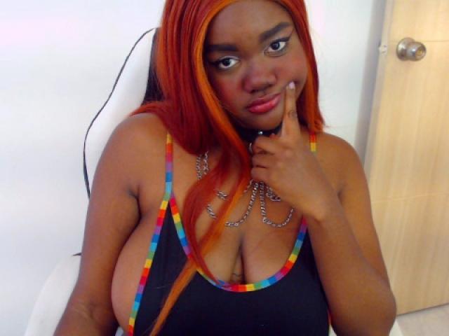 RoseBeckers has red hair, large tits, and is wearing a colorful, rainbow-trimmed black top with chains. She's pointing at her lips with a finger.