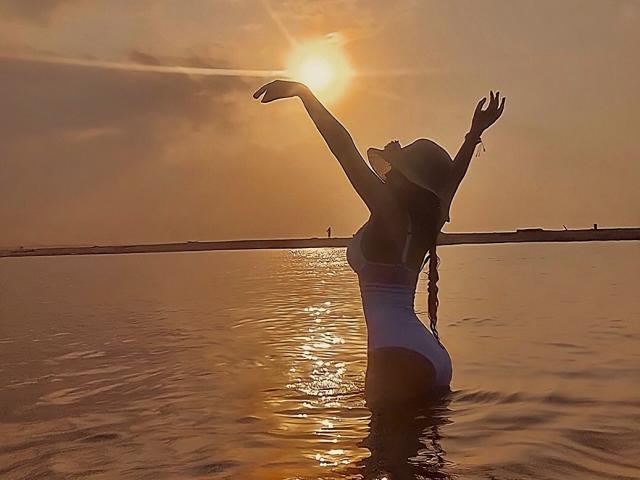 Silhouetted DanesaCronin in a white one-piece, arms raised, wearing a hat, standing in water at sunset with sun glowing behind her, reflecting off the surface.