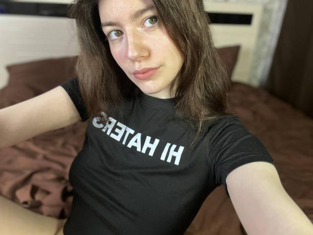 VinnaSkyHott mit braunen Haaren, die ein schwarzes T-Shirt trägt, sitzt auf einem Bett mit braunen Laken, zeigt ihre Brüste und lächelt leicht in die Kamera..