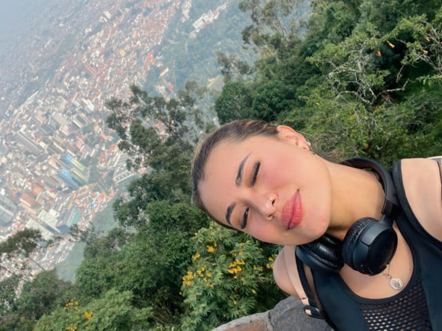 ArianaBellee with her brown hair in a bun, wearing black headphones, has her eyes closed and a slight smile, showing her pink nose and red lipstick. Her tits are small.