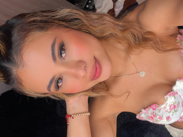 ArianaBellee has wavy blonde hair, brown eyes, and a nose ring. She's wearing a floral top, showing her tits, with a gold necklace and red bracelet.