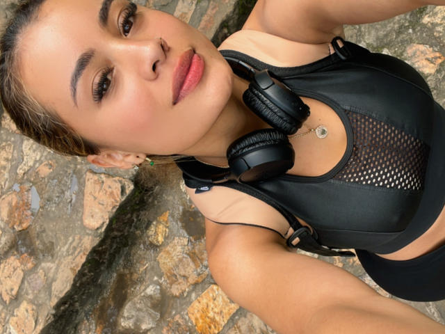 ArianaBellee with brown eyes, pink lips, and black mesh sports bra, wearing black headphones, has a small belly button piercing, and is looking at the camera.