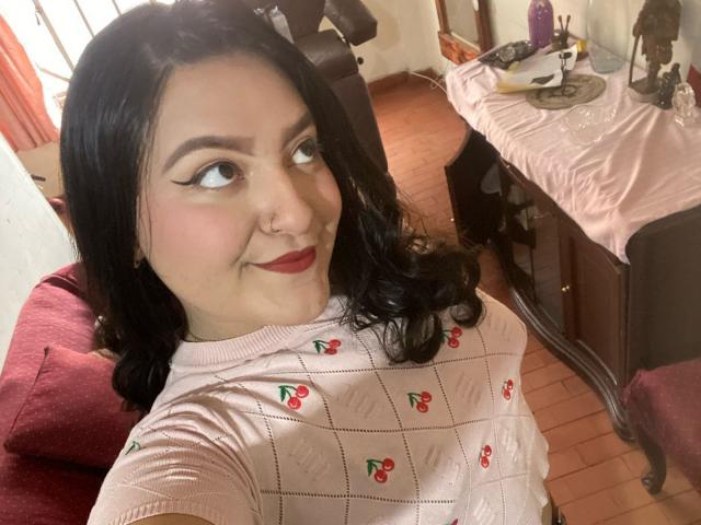 ShopiaRosse with black hair, red lipstick, and eyeliner, wearing a pink cherry-patterned sweater, smiling at the camera, sitting on a red cushion, wooden floor.
