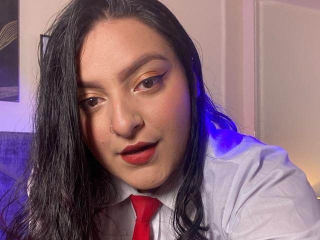 ShopiaRosse has long black hair, a nose ring, and is wearing a white shirt with a red tie. She has red lipstick and blue light on her right.