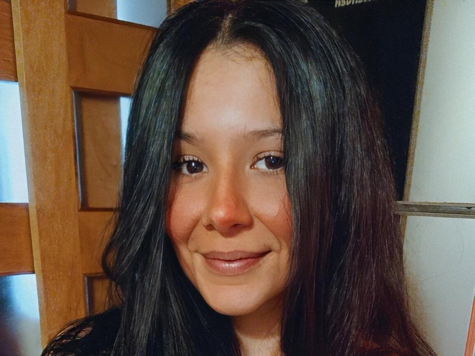 LissaThompron has straight black hair, brown eyes, and a small smile with a slight blush on her cheeks. She's wearing a black top, and the wood chair is visible.