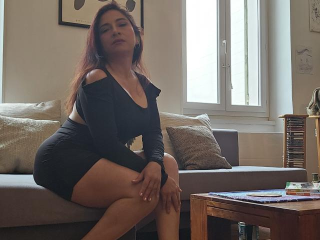 AlessiaRed, con su largo cabello rojo, está sentada en un sofá gris, vestida con un mini-vestido negro de manga larga y escote pronunciado, que deja al descubierto sus pechos y glúteos, en una habitación iluminada por el sol..