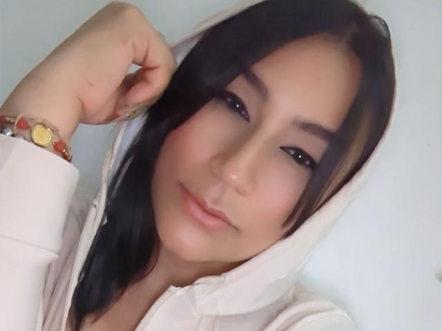 EmaFoxy with black hair, wearing a white hoodie, has her hand near her face, smiling slightly, wearing a red and gold bracelet, and has subtle makeup.