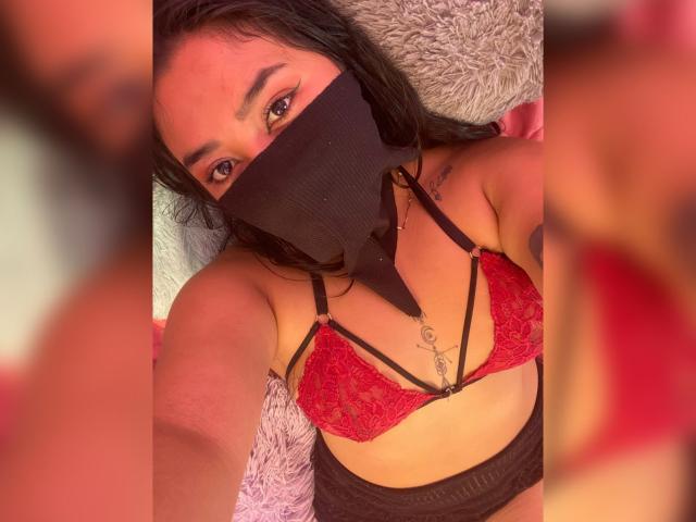 Alana Halamm mit dunklem Haar, trägt einen roten Spitzen-BH und eine schwarze Maske, zeigt ihre Brüste und schwarze Unterwäsche und liegt auf einem flauschigen Kissen. Ihre Augen sind sichtbar..
