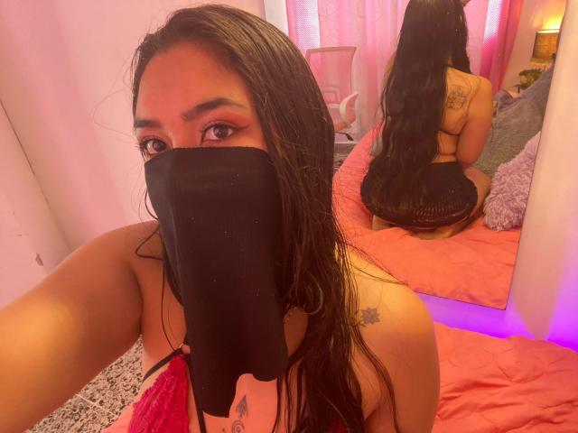 AlanaHalamm con el cabello negro y mojado, lencería rosa, máscara negra, tatuajes visibles, sentada en una cama rosa, de espaldas a la cámara, mostrando su trasero, reflejo en un espejo, habitación rosa, luz púrpura..