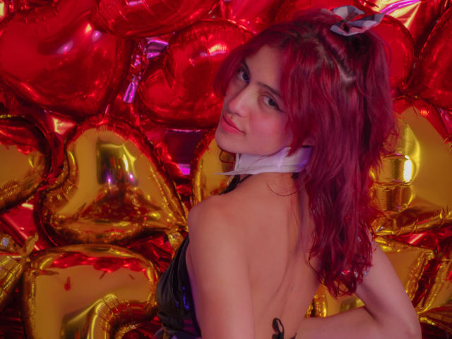 CameronIggy69 con cabello rojo, cinta blanca alrededor del cuello, top negro brillante, de espaldas a la cámara, rodeada de globos rojos y dorados, mirando por encima del hombro, sonriendo..