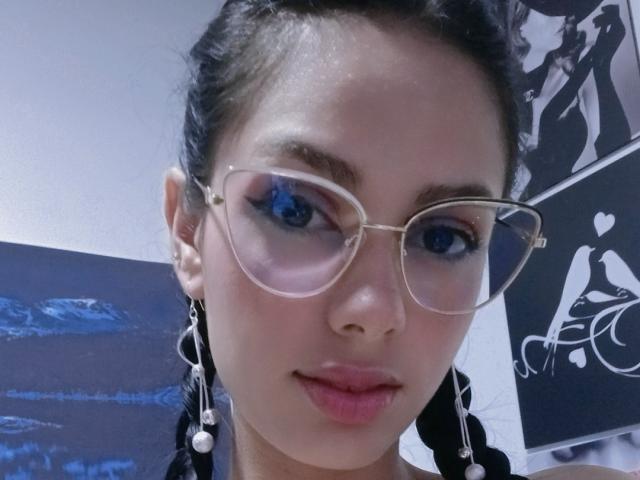 AnaisHilton with clear glasses, blue eyes, and black braided pigtails, showing her pouty lips and wearing long pearl earrings. Her tits are small and she's looking at the camera.