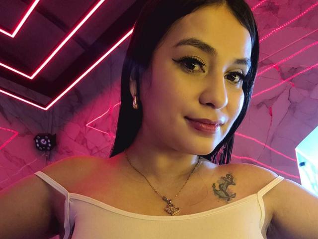 AliceNova with black hair, eyeliner, and pink lipstick, wearing a pink top, showing a tattoo of an anchor on her right shoulder, neon lights in the background.