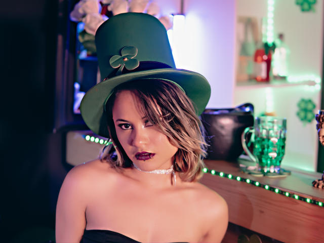 CassieJones with green top hat, dark lipstick, and choker, showing off her bare tits, short blonde hair, and a sexy look in a bar with green LED lights.