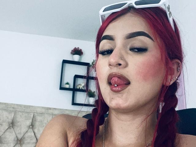 xHottieBabeX con trenzas rojas, piercing en la lengua y maquillaje, sacando la lengua, usando gafas de sol blancas, sonrojándose y mostrando su piercing en la nariz. Sus pechos son ligeramente visibles..