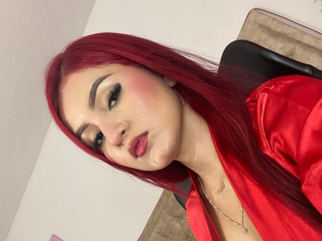 xHottieBabeX met rood haar, dikke wenkbrauwen, roze blush, zwarte eyeliner, rode lipstick, een neusring en een rode satijnen blouse, kijkt naar beneden met licht geopende rode lippen..