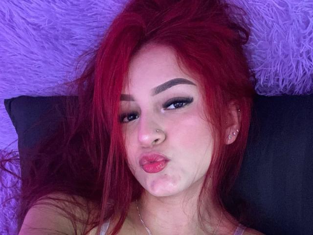 xHottieBabeX com cabelo ruivo, lábios rosados e carnudos, delineador preto, piercing no nariz e um pequeno hematoma nos lábios, deitada em um travesseiro preto, com iluminação roxa..