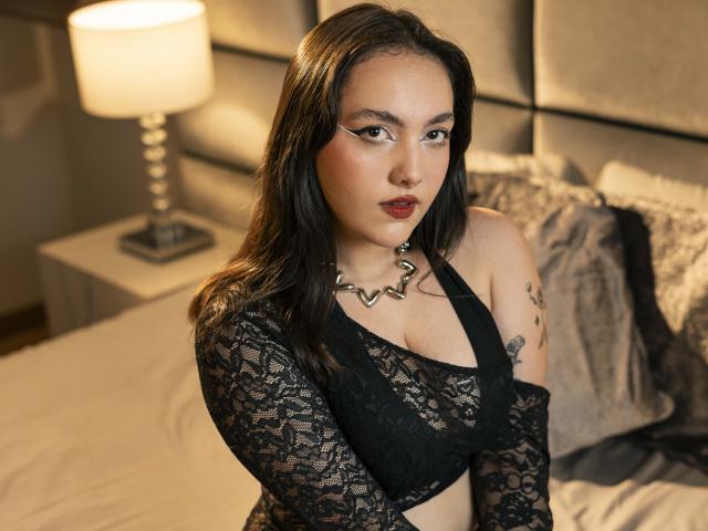 TabitaMaddoxx has dark hair, black lace top showing her tits, tattooed arm, and a silver necklace, looking seductively at the camera with red lipstick. Bedroom setting.