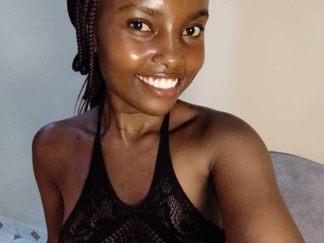Africanah with dark brown skin, smiling, wearing a black mesh halter top, showing her tits, braided hair, and shiny skin, in a selfie.