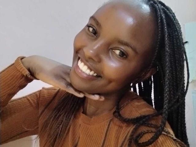 Africanah with dark brown skin, smiling, wearing a ribbed, brown, long-sleeve top, and braided hair, resting her chin on her hand. Her teeth are visible.