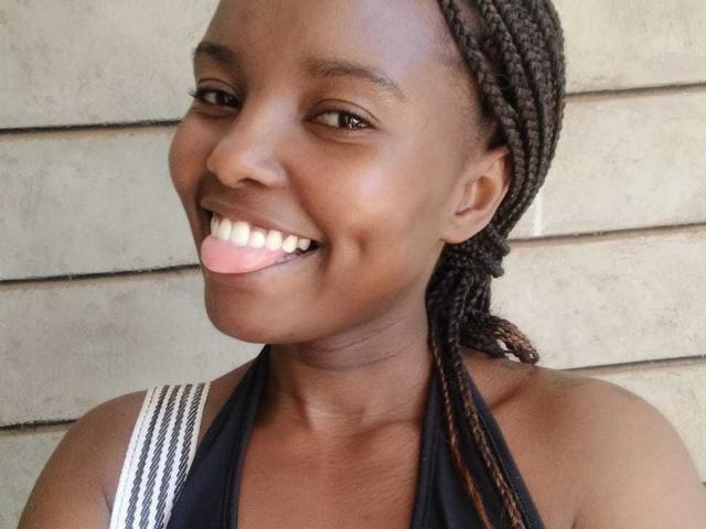 Africanah, smiling with tongue out, dark skin, braided hair, black halter top, showing teeth, brown eyes, sleeveless, concrete wall in background, upper body shot, cute.