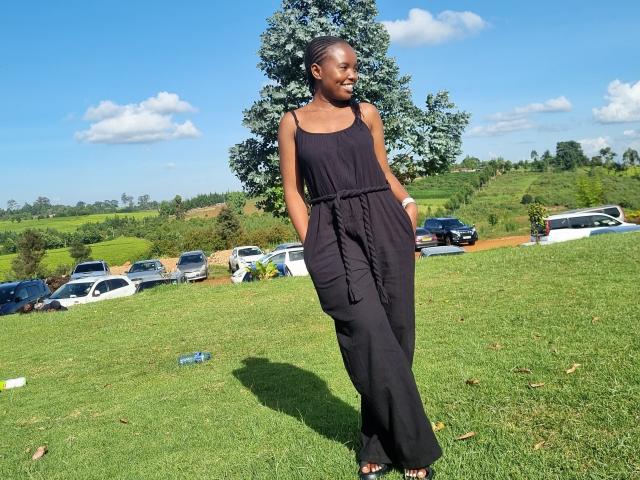 Africanah stands on grass wearing a black sleeveless dress with hands in pockets, smiling, dark skin, short hair, small tits, no visible ass, blue sky, scattered clouds, parked cars.