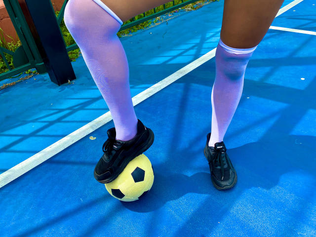 SophieSonyas' legs in white, sheer, thigh-high socks standing on a blue, textured, outdoor court with black sneakers on a soccer ball, sunlight casting shadows.