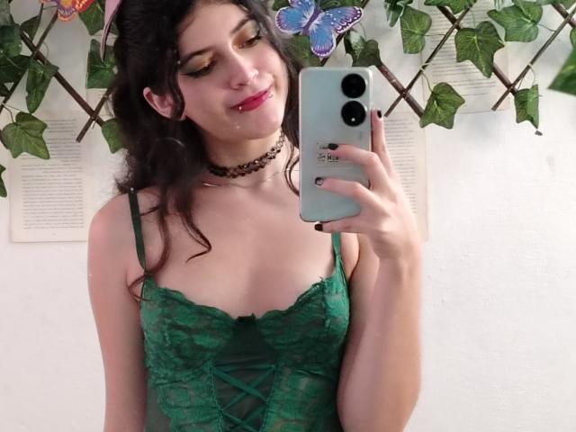 RachelHottie with wavy dark hair, green lace lingerie, choker, holding phone, smiling, pink lipstick, butterfly decor, white wall, green leaf vines, slightly visible boobs.