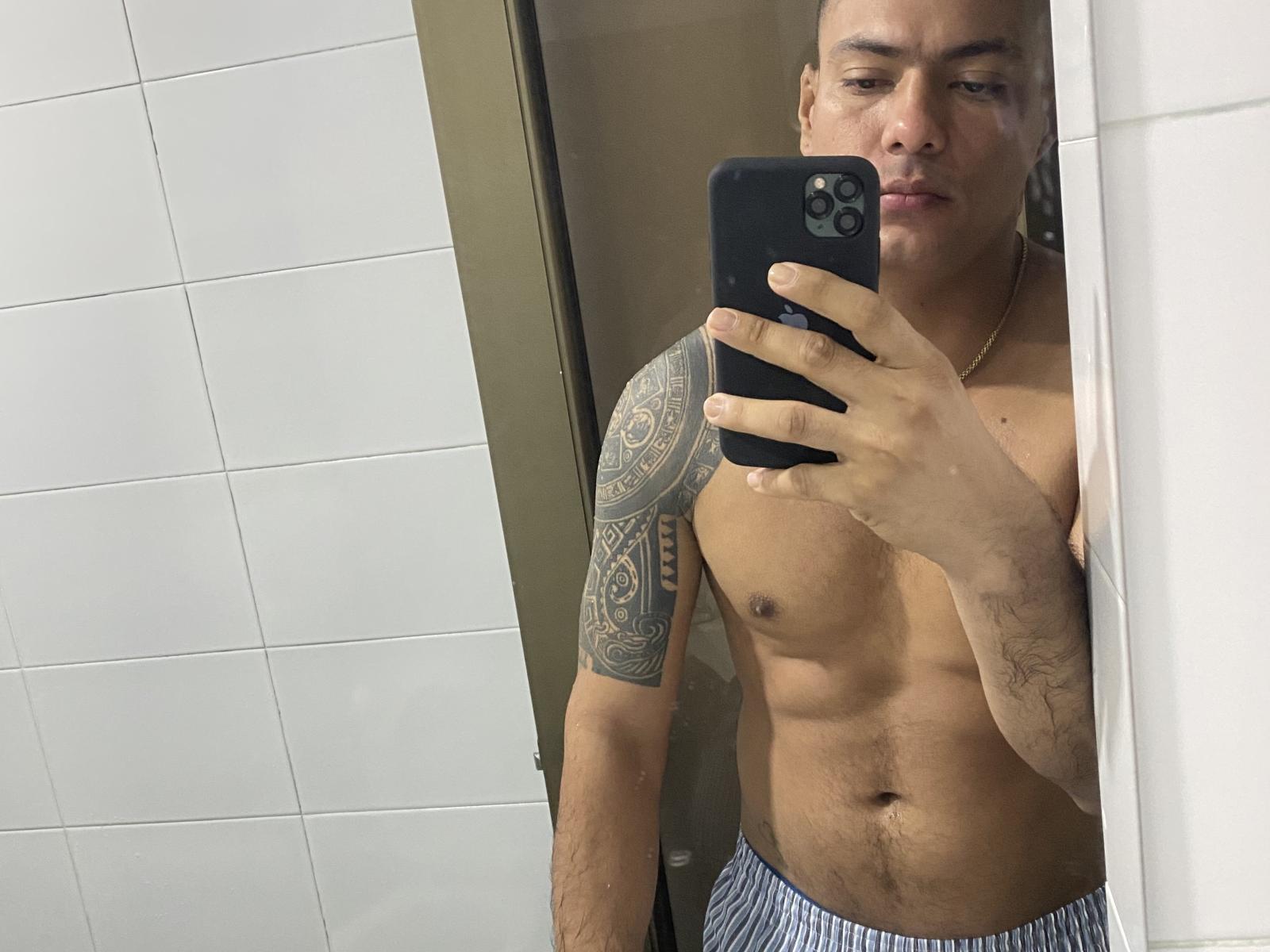 LeonHard, shirtless, holding phone in mirror selfie, tattooed arm, muscular, tan skin, short hair, wearing blue-striped pants, serious expression, gold necklace, reflection shows white tiled wall.