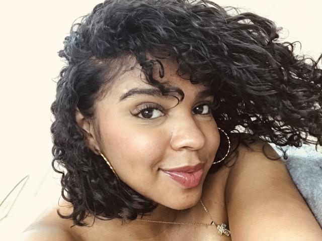 LisaMendez has curly black hair, brown eyes, and a light brown skin tone. She's sticking her tongue out, wearing gold hoop earrings, and a necklace. Boobs visible.
