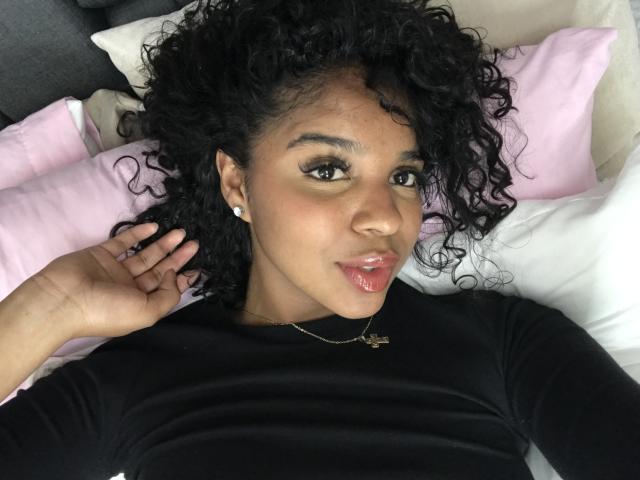 LisaMendez with curly black hair, brown skin, and glossy pink lips, wearing a black top, lying on pink and white pillows, showing a hand near her face.