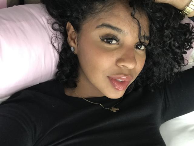 LisaMendez with curly black hair, wearing a black shirt, lying on a pink pillow, has shiny pink lips, dark eyeliner, and a gold necklace with a star pendant.