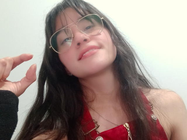MaddieHottie with long dark hair, glasses, and a red halter top, showing her cute smile and slightly raised head. She's touching her hair with her right hand.