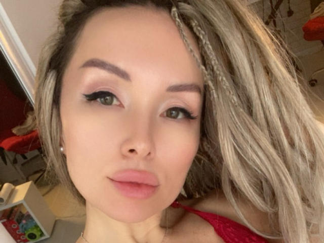 AvaGreenx with blonde braids, green eyes, and pink lips, wearing red lingerie, has full, puffy tits, and a slightly upturned nose, looking directly at the camera.