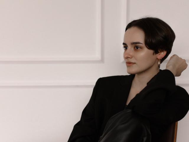 BellaSpace with short brown hair, black outfit, and earrings, sitting with black leather pants, arm resting on chair, white wall in background, slight smile, looking to the side.