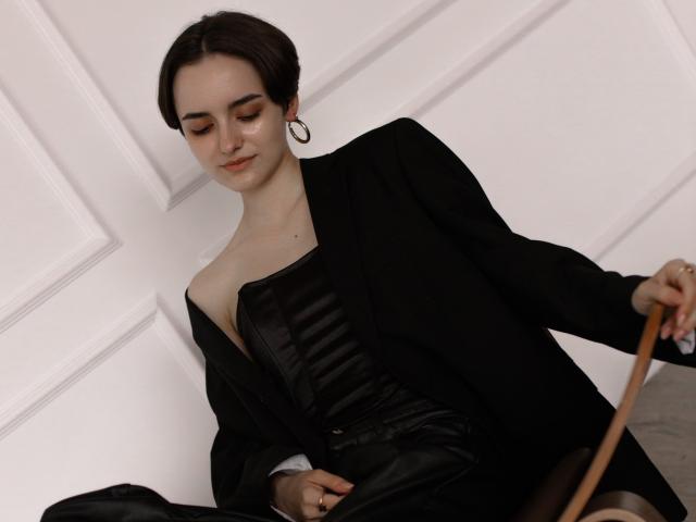 BellaSpace with short brown hair, hoop earrings, black strapless corset, and black jacket, holding a wooden stick, sitting against white walls, looking down with a subtle smile.
