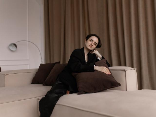 BellaSpace with short brown hair, black outfit, and black boots, leans on beige sofa, arms resting on brown pillows, looking at camera with a relaxed, slightly bored expression.