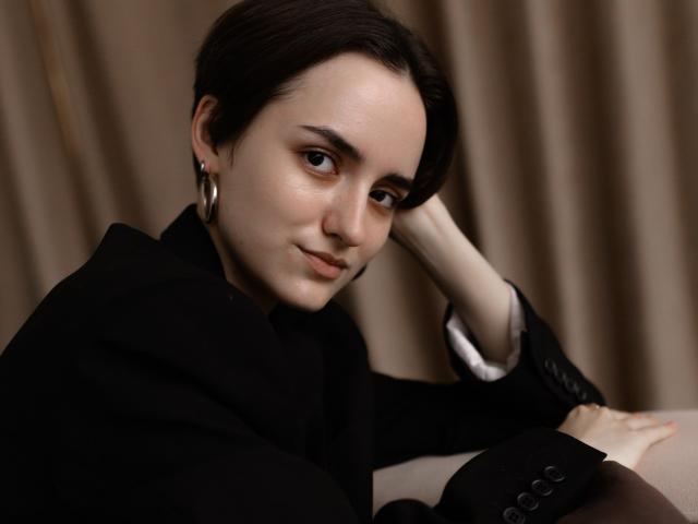 BellaSpace has short dark hair, a cute face, and small tits, wearing a black outfit with silver hoop earrings, resting her head on her hand, looking slightly to the side.