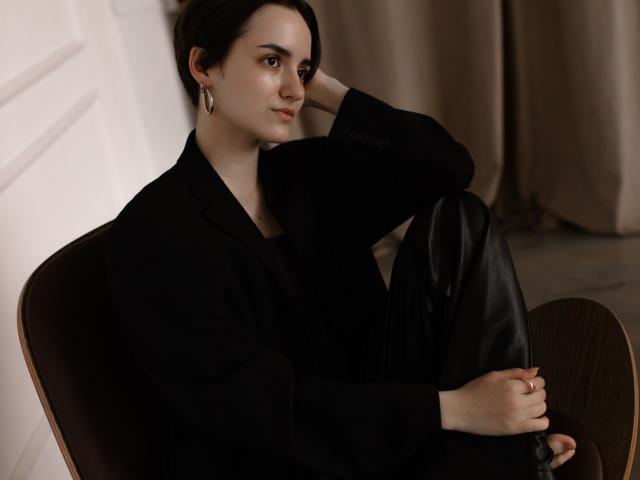 BellaSpace with short black hair, wearing a black blazer and leather pants, sits in a wooden chair, hand on knee, looking to the side with a slight smile.
