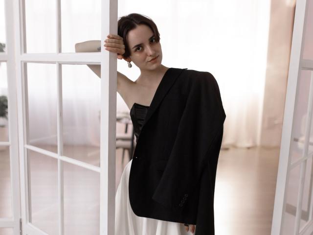BellaSpace with dark brown hair, black jacket, and white dress, standing in a doorway, leaning on a white frame, looking at the camera, in a soft, natural light.