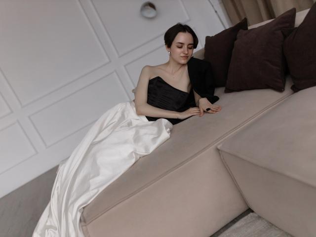 BellaSpace with small tits, wearing a black strapless top and white satin skirt, sitting on a beige couch, touching a brown pillow, in a softly lit room.