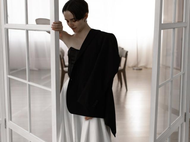 BellaSpace with dark hair, black shawl, and white dress, standing in a doorway, holding door frame, looking down, bare shoulders, subtle makeup, soft light, elegant.