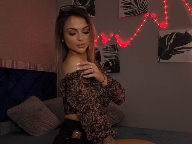 KarolinaSykes with blonde hair, wearing an off-shoulder brown patterned top, black shorts, red string lights, and sunglasses on her head, touching her shoulder, in a dimly lit room.