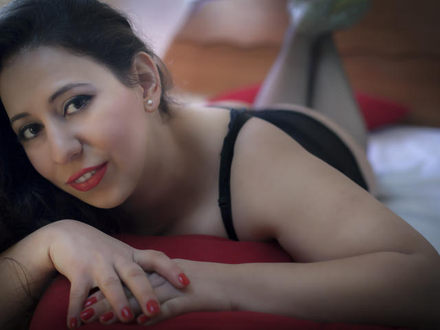 RenattaRosse liegt auf einem Bett, trägt einen schwarzen BH, hat rote Nägel, lächelt, hat dunkles Haar, blasse Haut, Perlenohrringe, ihre Hände liegen auf einem roten Kissen, die Beine sind angewinkelt, sie trägt schwarze Unterwäsche, das Licht ist weich, die Atmosphäre ist intim..